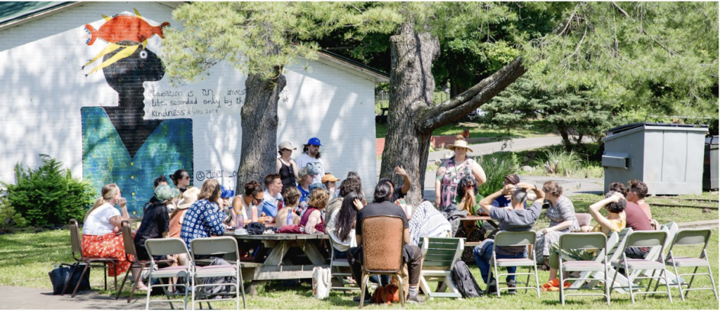 Masked people sitting in a circle outside of a white house and large tree.